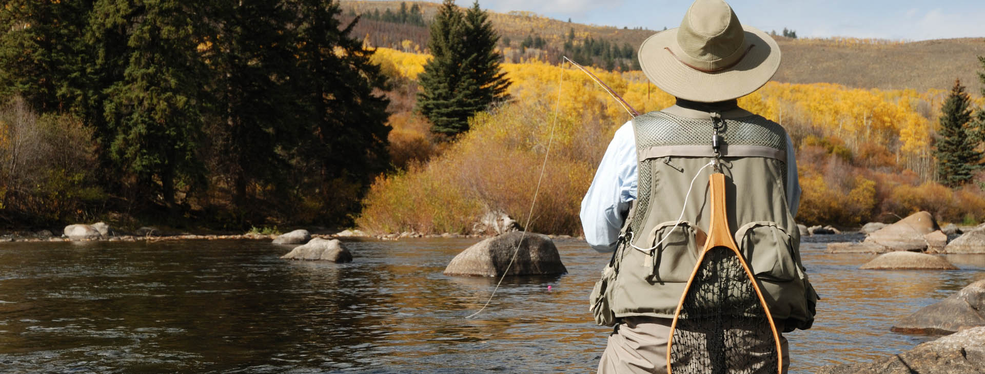 A man wearing a hat and vest stands on a rock in the river, surrounded by flowing water and natural scenery.