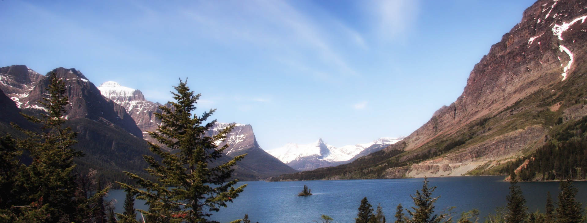 A serene lake bordered by majestic mountains and lush green trees under a clear blue sky.