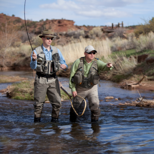 Two men are fishing together in a serene river, surrounded by lush greenery and calm waters.