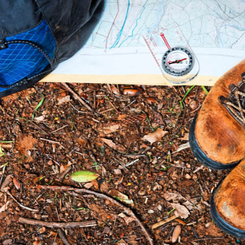 Hiking boots, a map, and a compass arranged on the ground, indicating preparation for an outdoor adventure