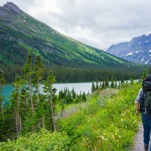 A man with a backpack walks along a mountain trail, surrounded by lush greenery and rocky terrain